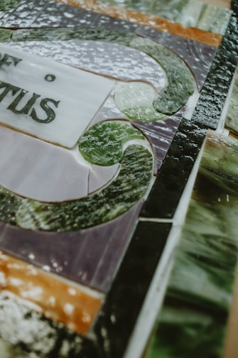 A close-up of a stained glass artwork with intricate patterns and text. The image features a mix of smooth and textured glass pieces in various colors, arranged in a detailed design.