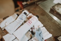 Several tubes of ink or paint are laid out on a wooden surface. The tubes are mostly white with some text and logos in colors like blue and red. Some tubes are open and appear used, with traces of ink around them. A piece of textured cloth or paper is also present next to the tubes.