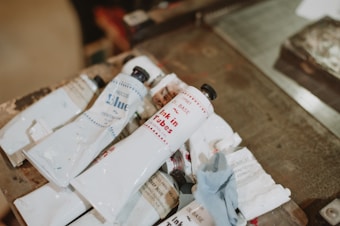 Several tubes of ink or paint are laid out on a wooden surface. The tubes are mostly white with some text and logos in colors like blue and red. Some tubes are open and appear used, with traces of ink around them. A piece of textured cloth or paper is also present next to the tubes.
