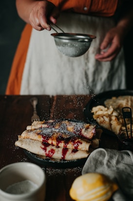 A person is sprinkling powdered sugar over freshly made crepes filled with a red berry sauce. The scene is set on a wooden table with a rustic feel, accompanied by a lemon, a small bowl, a jar, and other kitchen utensils.