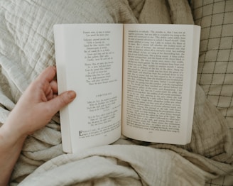A close-up view of an open book being held by a hand against a soft, textured fabric background. The book is open to a page with English text that includes a chapter heading. The hand holding the book is partially visible, with neatly trimmed nails.