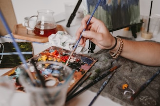A hand holding a paintbrush over a palette filled with various vibrant colors on a table. The scene includes additional paintbrushes in a container, a cloth, a glass teapot on a wooden tray, and a partially painted canvas. The atmosphere is creative and artistic.