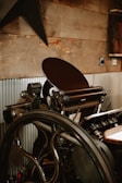 A vintage printing press with a large wheel and intricate mechanical components is positioned against a rustic wooden wall. The press has various metallic parts, handles, and rollers, with corrugated metal sheets partially covering the lower wall section.