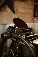 A vintage printing press with a large wheel and intricate mechanical components is positioned against a rustic wooden wall. The press has various metallic parts, handles, and rollers, with corrugated metal sheets partially covering the lower wall section.