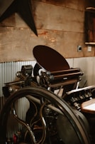 A vintage printing press with a large wheel and intricate mechanical components is positioned against a rustic wooden wall. The press has various metallic parts, handles, and rollers, with corrugated metal sheets partially covering the lower wall section.