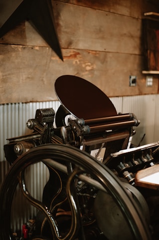 A vintage printing press with a large wheel and intricate mechanical components is positioned against a rustic wooden wall. The press has various metallic parts, handles, and rollers, with corrugated metal sheets partially covering the lower wall section.
