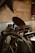 A vintage printing press with a large wheel and intricate mechanical components is positioned against a rustic wooden wall. The press has various metallic parts, handles, and rollers, with corrugated metal sheets partially covering the lower wall section.