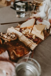 A wooden platter is covered with an assortment of cheeses, charcuterie, and nuts. There are several types of cheese, including wedge shapes and soft rounds. Crackers are scattered among the cheese, and there are nuts such as pistachios visibly placed around the platter. In the background, there are clear glasses containing water.