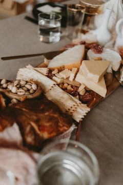 A wooden platter is covered with an assortment of cheeses, charcuterie, and nuts. There are several types of cheese, including wedge shapes and soft rounds. Crackers are scattered among the cheese, and there are nuts such as pistachios visibly placed around the platter. In the background, there are clear glasses containing water.