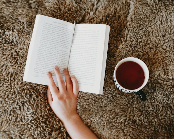 An open book is placed on a fluffy, textured rug. A hand is resting on the book, suggesting someone is reading. A cup filled with a dark beverage, likely tea or coffee, is positioned nearby. The scene appears cozy and inviting.