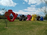 Large, colorful sculptures shaped like the word 'Oaxaca' are arranged in an open grassy area, surrounded by sparse trees. The sky above is blue with scattered clouds, creating a vibrant and lively atmosphere. Mountains are visible in the background.