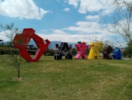 Large, colorful sculptures shaped like the word 'Oaxaca' are arranged in an open grassy area, surrounded by sparse trees. The sky above is blue with scattered clouds, creating a vibrant and lively atmosphere. Mountains are visible in the background.