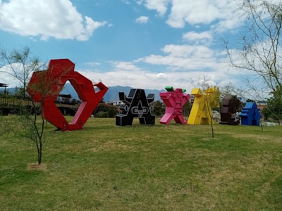 Large, colorful sculptures shaped like the word 'Oaxaca' are arranged in an open grassy area, surrounded by sparse trees. The sky above is blue with scattered clouds, creating a vibrant and lively atmosphere. Mountains are visible in the background.