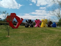 Large, colorful sculptures shaped like the word 'Oaxaca' are arranged in an open grassy area, surrounded by sparse trees. The sky above is blue with scattered clouds, creating a vibrant and lively atmosphere. Mountains are visible in the background.