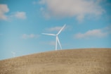 A landscape featuring open fields under a clear blue sky with scattered clouds. Two wind turbines stand prominently on a rolling hill, their blades adding a sense of rotation and movement. The scene conveys a sense of natural openness and renewable energy.