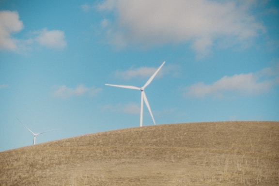 A landscape featuring open fields under a clear blue sky with scattered clouds. Two wind turbines stand prominently on a rolling hill, their blades adding a sense of rotation and movement. The scene conveys a sense of natural openness and renewable energy.