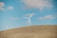 A landscape featuring open fields under a clear blue sky with scattered clouds. Two wind turbines stand prominently on a rolling hill, their blades adding a sense of rotation and movement. The scene conveys a sense of natural openness and renewable energy.