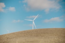 A landscape featuring open fields under a clear blue sky with scattered clouds. Two wind turbines stand prominently on a rolling hill, their blades adding a sense of rotation and movement. The scene conveys a sense of natural openness and renewable energy.