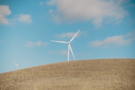 A landscape featuring open fields under a clear blue sky with scattered clouds. Two wind turbines stand prominently on a rolling hill, their blades adding a sense of rotation and movement. The scene conveys a sense of natural openness and renewable energy.
