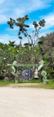 A large gong is suspended between two intricately carved statues of mythical or legendary creatures, possibly in a traditional or cultural setting. The surrounding landscape is lush with greenery, featuring various trees and foliage under a bright blue sky with scattered clouds.