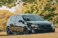 A sleek black Volkswagen GTI is parked on a grassy field, surrounded by lush green trees under a clear sky. The car's glossy finish reflects its surroundings, and it features tinted windows. A large decal with Japanese characters is visible on the front windshield.
