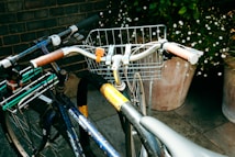 Two bicycles are parked close to each other, featuring baskets attached to the handlebars. One of the bikes has a worn and rusted frame, with a drink can placed in its rear rack. The setting includes potted plants with small white flowers and a brick wall backdrop, casting natural light and shadows on the scene.