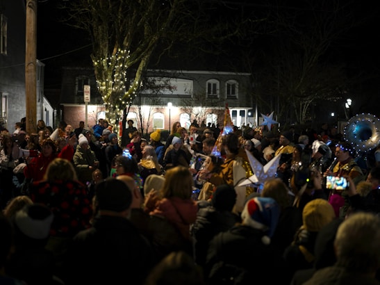 A large crowd gathers outdoors during the nighttime, many wearing festive hats and attire. Several people hold decorative stars, and the scene is lit by string lights wrapped around a tree. A sense of celebration and community is conveyed by the crowd's engagement and proximity.