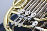 A close-up view of a brass musical instrument, specifically a French horn, highlighting the intricate valves and tubing. The metal parts are polished and shiny, reflecting light, with a focus on the keys and moving parts that control the instrument's pitch.
