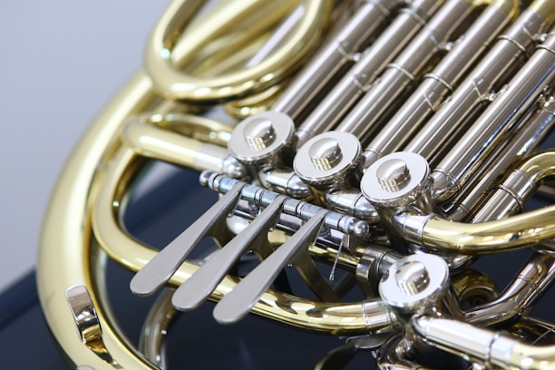 A close-up view of a brass musical instrument, specifically a French horn, highlighting the intricate valves and tubing. The metal parts are polished and shiny, reflecting light, with a focus on the keys and moving parts that control the instrument's pitch.