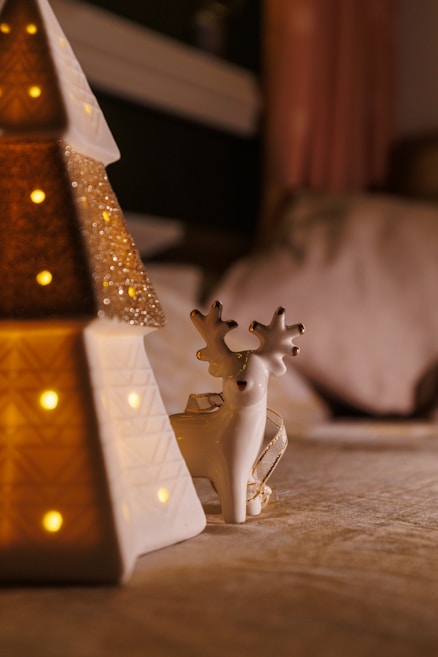 A ceramic reindeer figurine with brown antlers and a gold ribbon stands on a surface next to a lit-up decorative ceramic Christmas tree with small lights embedded in it. The scene is set indoors with warm lighting, creating a cozy ambiance.
