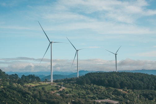 Three large wind turbines stand prominently against a backdrop of rolling hills and dense green forests. The sky is expansive, with scattered clouds and a soft blue hue.