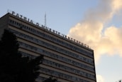 A multi-story building with a message in Spanish on its rooftop, set against a sky with clouds. The building has numerous windows and appears to have air conditioning units attached to some of them.