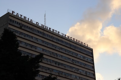 A multi-story building with a message in Spanish on its rooftop, set against a sky with clouds. The building has numerous windows and appears to have air conditioning units attached to some of them.