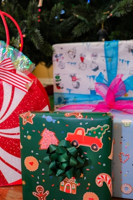 Several colorful Christmas presents are placed under a Christmas tree. One gift is wrapped in festive green paper adorned with holiday-themed images such as gingerbread men, cookies, a red truck carrying a Christmas tree, and candy canes. It is topped with a green bow. Another present is wrapped in light blue paper with whimsical winter graphics and features a pink bow. The red and white round gift bag has a peppermint design with shiny, iridescent wrapping at the top. The background is partially obscured by the dark green branches of the Christmas tree decorated with small string lights.