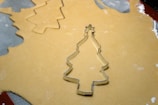 A metal Christmas tree-shaped cookie cutter is placed on rolled-out cookie dough alongside other cut-out tree shapes. The dough is laid on a surface with a mesh pattern visible in the background, and some flour is scattered around.