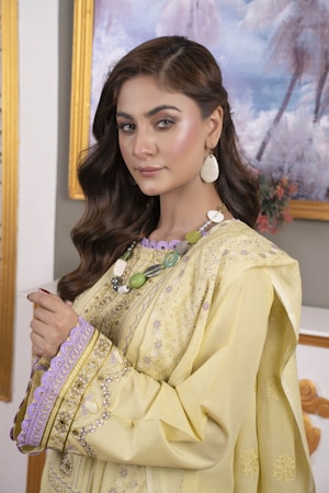 A woman is wearing a yellow traditional dress with intricate embroidery and lacework. She has long, wavy brown hair and is wearing large, oval earrings. The background features a framed painting with a soft palette of blues and purples.