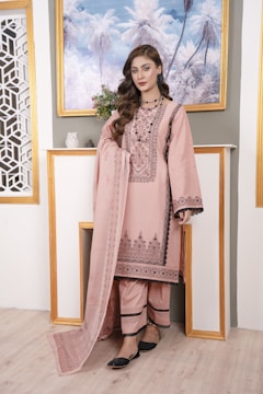 A woman is standing indoors wearing a traditional pink outfit with intricate patterns. She is posing in front of a decorative background with a painting of palm trees and a shelf with small decorative items. Her hair is styled in loose waves and she is wearing black shoes.