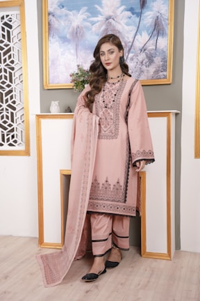 A model wearing a pastel pink suit with traditional motifs, standing in a sunlit room.