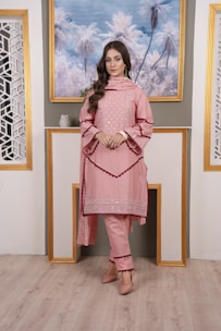 A woman is standing indoors in a stylish outfit. She is wearing a pink embroidered kurta with matching pants and a dupatta. The ensemble has intricate detailing around the borders. The setting includes a portrait of palm trees framed in gold on the wall, flanked by decorative lattice panels.
