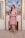 A woman is standing indoors in a stylish outfit. She is wearing a pink embroidered kurta with matching pants and a dupatta. The ensemble has intricate detailing around the borders. The setting includes a portrait of palm trees framed in gold on the wall, flanked by decorative lattice panels.