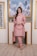 A woman is standing indoors in a stylish outfit. She is wearing a pink embroidered kurta with matching pants and a dupatta. The ensemble has intricate detailing around the borders. The setting includes a portrait of palm trees framed in gold on the wall, flanked by decorative lattice panels.