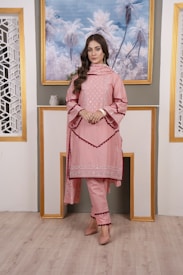 A woman is standing indoors in a stylish outfit. She is wearing a pink embroidered kurta with matching pants and a dupatta. The ensemble has intricate detailing around the borders. The setting includes a portrait of palm trees framed in gold on the wall, flanked by decorative lattice panels.