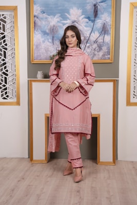 A woman is standing indoors in a stylish outfit. She is wearing a pink embroidered kurta with matching pants and a dupatta. The ensemble has intricate detailing around the borders. The setting includes a portrait of palm trees framed in gold on the wall, flanked by decorative lattice panels.