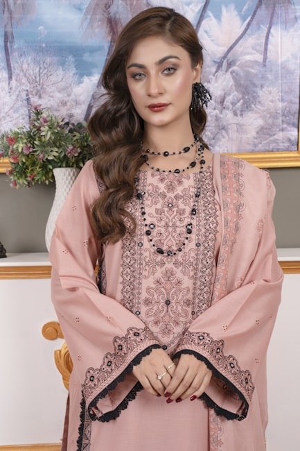 A woman in traditional attire with intricate black embroidery on a pale pink fabric. She is wearing a long, patterned dress complemented by an embroidered shawl draped over her shoulders. Her hair is styled in loose curls, and she accessorizes with black beaded jewelry and a floral hair accessory. She stands in front of a backdrop that features a serene painting of a snow-covered forest, with a white vase of flowers placed on a shelf beside her.