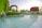 A garden scene featuring a lush, well-maintained landscape with a large, inviting pool in the foreground. The water is clear, with gentle ripples, and a small waterfall cascading from the side of the pool. In the background, there are several palm trees and hedges arching over pathways, adorned with vibrant pink flowers. The sky is clear, suggesting a calm, serene environment.