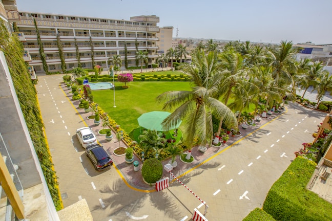 A large courtyard with well-maintained green grass and palm trees. There is a wide paved pathway surrounding the grassy area, with parking lines and two cars parked parallel. The background features a multi-story building with balconies adorned with potted plants, suggesting an organized and scenic environment.