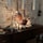 A cozy interior scene features a rustic wooden sideboard against a stone wall. On the sideboard, several lit candles create a warm and inviting ambiance. Decor includes a small decorative Christmas tree, dried cotton branches in a white vase, and scattered greenery. The overall setting is accentuated by hanging pendant lights and earthy tones.