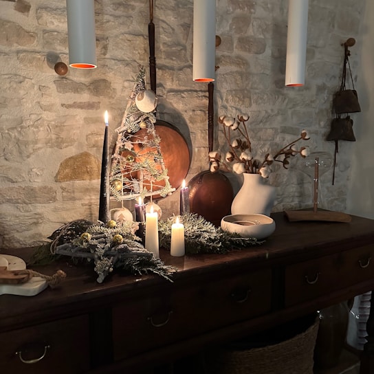 A cozy interior scene features a rustic wooden sideboard against a stone wall. On the sideboard, several lit candles create a warm and inviting ambiance. Decor includes a small decorative Christmas tree, dried cotton branches in a white vase, and scattered greenery. The overall setting is accentuated by hanging pendant lights and earthy tones.