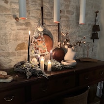A cozy interior scene features a rustic wooden sideboard against a stone wall. On the sideboard, several lit candles create a warm and inviting ambiance. Decor includes a small decorative Christmas tree, dried cotton branches in a white vase, and scattered greenery. The overall setting is accentuated by hanging pendant lights and earthy tones.
