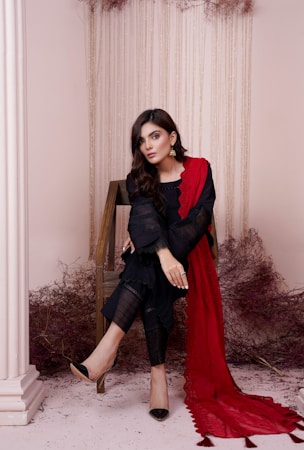 A woman sits elegantly on a wooden chair wearing a black outfit with intricate lace details, complemented by a flowing red scarf. Her hair is styled in loose waves and she wears gold earrings, exuding a sophisticated look. The setting includes creamy white pillars and a background draped with delicate, hanging decorative elements.